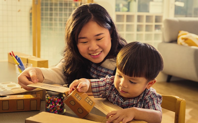 A teacher smiling while guiding a young child in a classroom activity, as the child plays with cardboard blocks and a jar of colourful beads