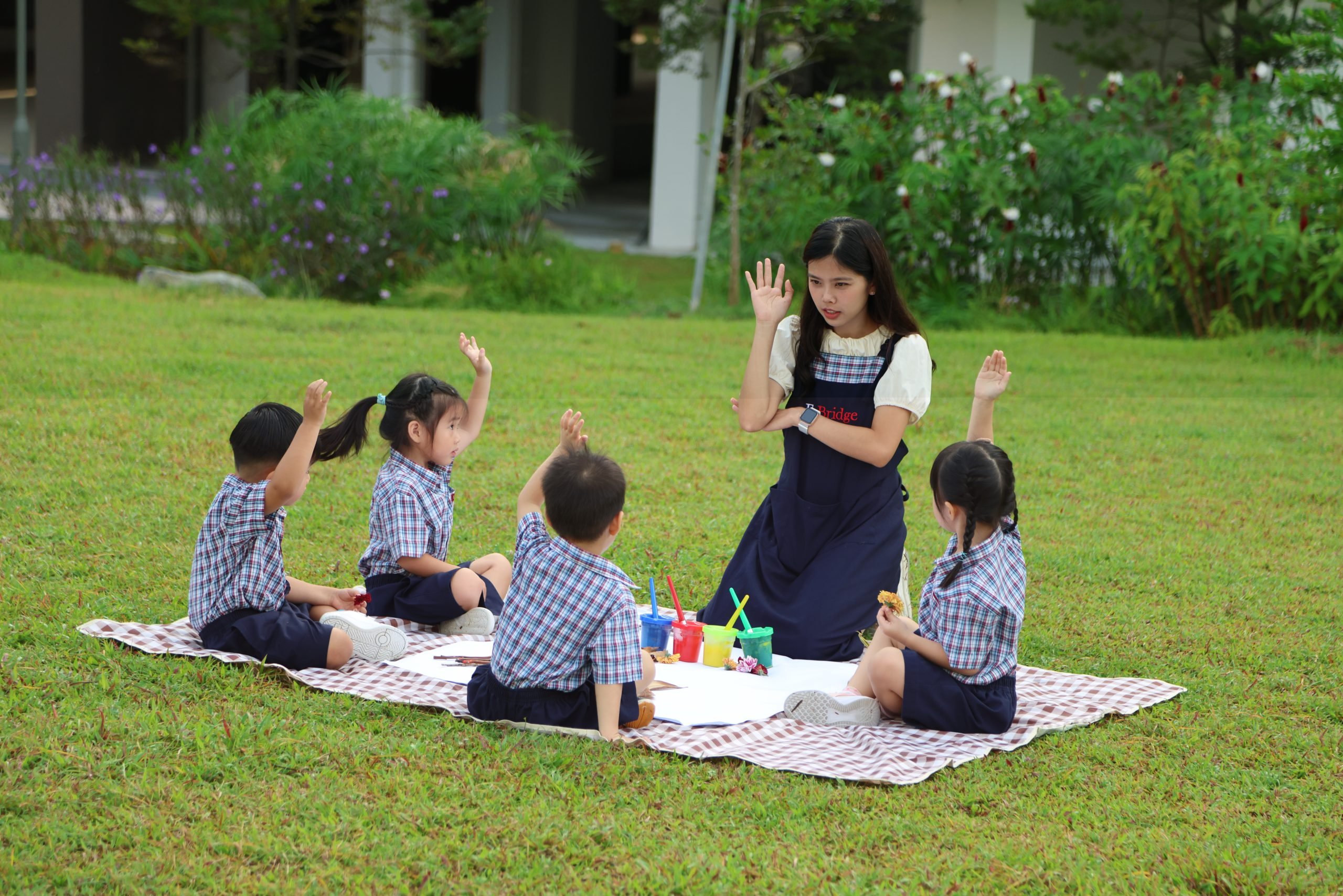 A teacher sitting on a picnic mat outdoors with four young children in school uniforms, engaging them in an interactive activity as they raise their hands