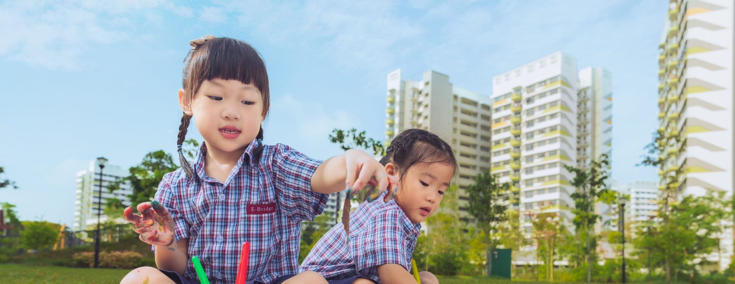 Two young children in school uniforms sitting on a picnic mat outdoors, engaging in a colorful finger-painting activity with brushes, paints, and paper.
