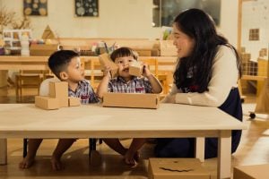 Two young children in school uniforms playing with cardboard blocks at a table, while a teacher sits beside them, smiling and engaging in the activity