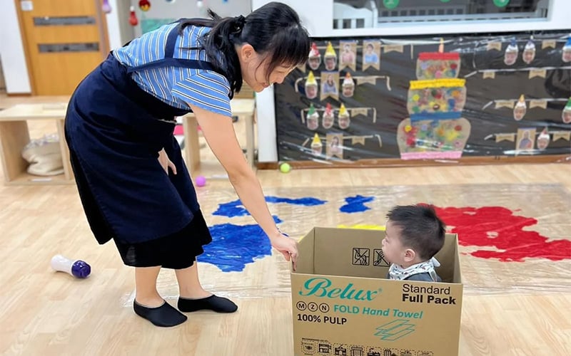 Teacher helping a young boy play in a cardboard box.