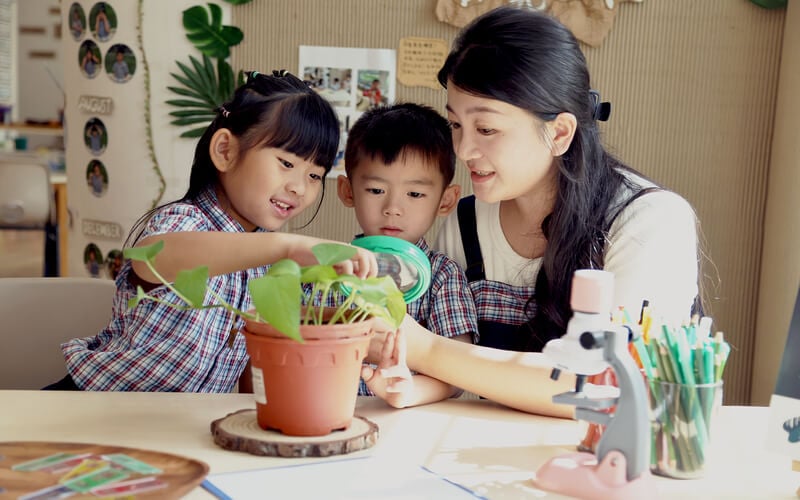 Teacher and children examining potted plant at Singapore preschool classroom.