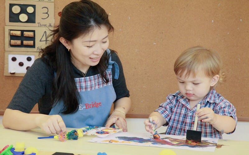 Teacher supporting young student during painting lesson at Singapore preschool